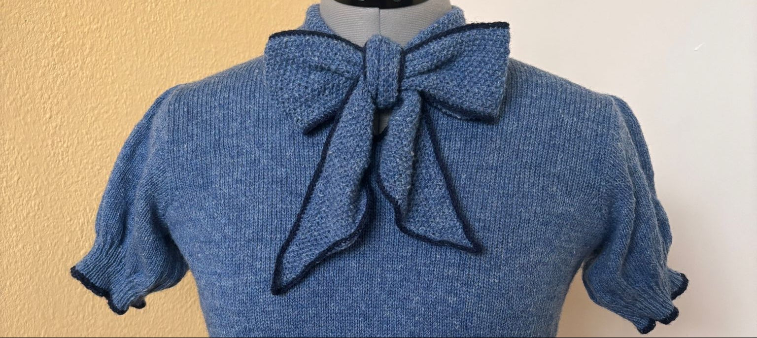 Blue knitted top with a bow on a mannequin against a yellow wall.
